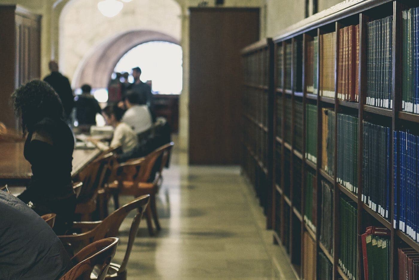 Readers at long tables beside tall shelves of bound volumes in a library