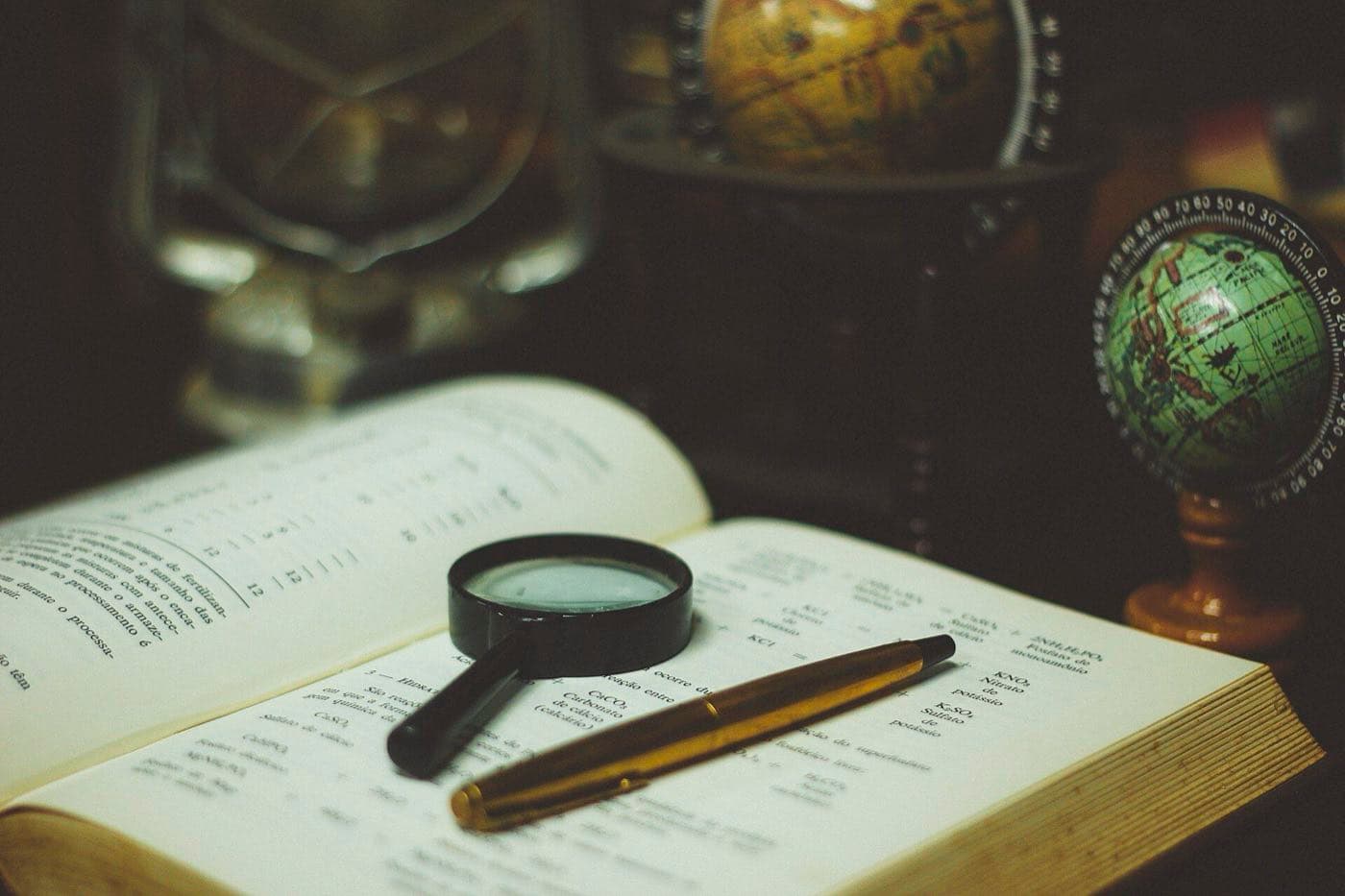 A magnifying glass and a pen resting on an open reference book beside two globes