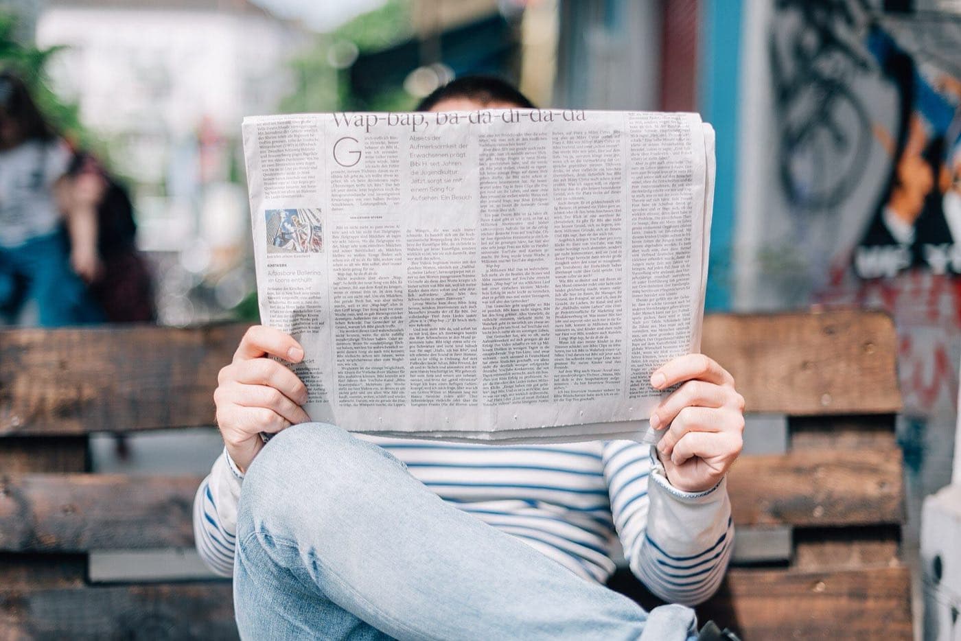 A person seated outdoors reading a broadsheet newspaper